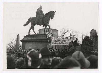 Demonstranten mit Plakat auf dem Karlsplatz, vermutlich vor dem Beginn der Rede von Karl Stetter. Auf dem Plakat ist zu lesen: „Wir sind gegen die Frankfurter Politik Ehrhardt [sic!] bringt: Not den Armen – Überfluß den Reichen“.