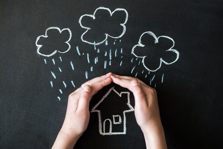 Ein Haus, über dem Wolken sich ausregnen. Schützend hält eine Person ihre Hände über das Haus.