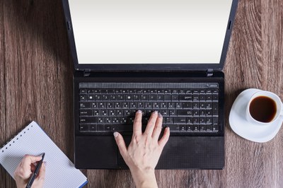 aufgeklappter Laptop, Bildschirm weiß; rechte Hand auf der Tastatur, linke hat Stift in der Hand; unter linker Hand Block mit karierten Seiten, unbeschrieben; rechts neben Tastatur Kaffeetasse mit Untersetzer